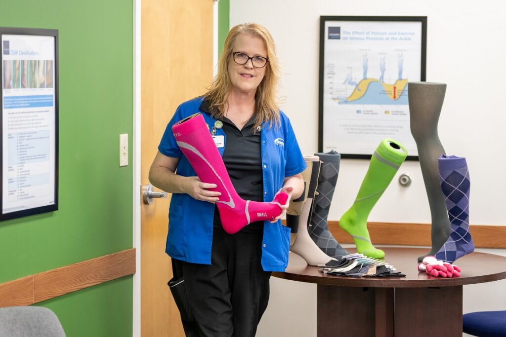 A compression fitter adjusting a colorful compression garment. The fitter is seen holding a garment. The background may feature medical equipment or shelves displaying compression garments. This image illustrates the process of fitting compression garments.